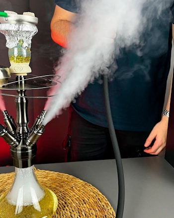 Kaljanas, stovėdamas ant apvalaus austo kilimėlio, rūksta dūmus. Kaljanas turi kelias žarnas, ir dūmai akivaizdžiai veržiasi iš kelių angų. Netoliese iš dalies matomas žmogus, laikantis žarną. Atmosfera rodo, kad kaljanas yra naudojamas, o dūmų debesys sukuria švelnų ir šiek tiek dramatišką vaizdą.
