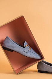 A pair of light blue suede loafers displayed with one shoe resting inside an open shoebox, creating an artistic composition. The box's interior is a contrasting shade of pink, set against a warm beige background.