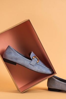 A pair of light blue suede loafers displayed with one shoe resting inside an open shoebox, creating an artistic composition. The box's interior is a contrasting shade of pink, set against a warm beige background.