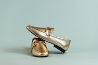 A pair of metallic gold loafers is placed elegantly against a soft blue background. One loafer is positioned on top of the other, showcasing its glossy finish and sleek design.