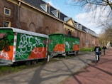 Two trailers with graffiti on them are parked alongside a brick building with arched windows and a decorative entrance. The trailers are primarily orange and green with various graffiti tags in different colors. Two people are riding bicycles on a path nearby on a sunny day.