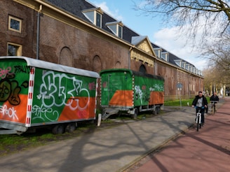 Two trailers with graffiti on them are parked alongside a brick building with arched windows and a decorative entrance. The trailers are primarily orange and green with various graffiti tags in different colors. Two people are riding bicycles on a path nearby on a sunny day.