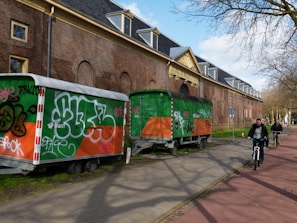 Two trailers with graffiti on them are parked alongside a brick building with arched windows and a decorative entrance. The trailers are primarily orange and green with various graffiti tags in different colors. Two people are riding bicycles on a path nearby on a sunny day.