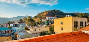 A scenic view of a small town with a cluster of buildings surrounded by lush green hills. The foreground features rooftops, some with solar panels, and trees dot the landscape. In the distance, a larger hill is visible under a clear blue sky.
