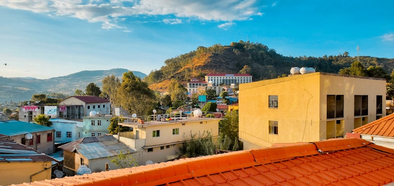 A scenic view of a small town with a cluster of buildings surrounded by lush green hills. The foreground features rooftops, some with solar panels, and trees dot the landscape. In the distance, a larger hill is visible under a clear blue sky.
