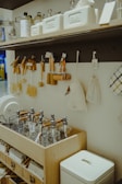 A wooden shelf and countertop display various kitchen and home items. The shelf holds glass jars, white containers, and cleaning supplies. Several wooden brushes, cloth bags, and kitchen tools hang from hooks. Below, there are glass dispensers with pumps neatly arranged in a wooden organizer.