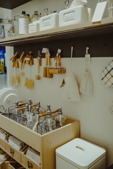 A wooden shelf and countertop display various kitchen and home items. The shelf holds glass jars, white containers, and cleaning supplies. Several wooden brushes, cloth bags, and kitchen tools hang from hooks. Below, there are glass dispensers with pumps neatly arranged in a wooden organizer.