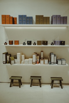Shelves display a variety of neatly arranged scented diffusers with elegant packaging in different colors. The top shelf holds boxes in hues of orange, blue, brown, and purple, while the lower shelf features diffusers and smaller products in matching tones. Below the shelves are three black wooden stools against a light-colored wall and floor.