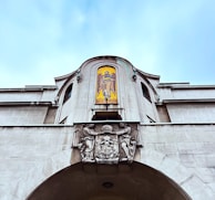 An ornate stone building featuring a carved crest with decorative elements below a colorful mosaic of an angel set into an arched niche. The sky is soft blue, providing a serene backdrop.