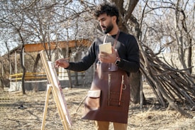 A person is painting outdoors in a natural setting with dry trees and a rustic backdrop. The individual is holding a brush and a palette, wearing a leather apron, and standing in front of an easel with a canvas. There is a partially built wooden structure and a pile of branches nearby.