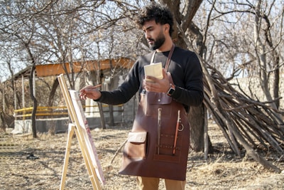 A person is painting outdoors in a natural setting with dry trees and a rustic backdrop. The individual is holding a brush and a palette, wearing a leather apron, and standing in front of an easel with a canvas. There is a partially built wooden structure and a pile of branches nearby.