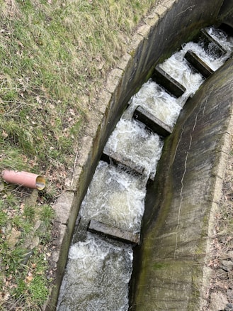 A small, structured water channel with concrete steps that facilitate the flow of water, surrounded by grassy banks. There is a red pipe on the left side near the edge of the channel.