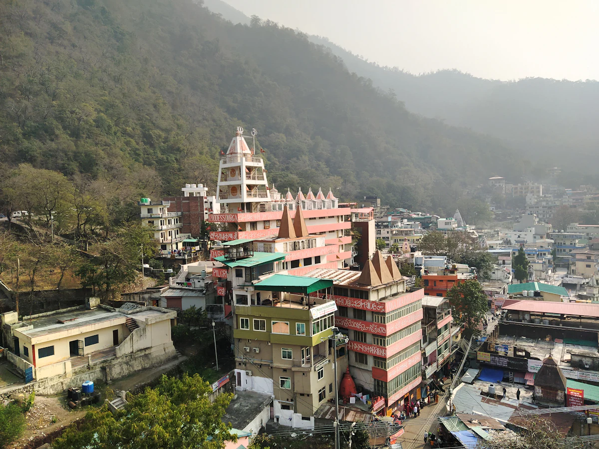 A tall tiered temple tower rising above hotels and shops on a forested hillside in the Laxman Jhula area of Rishikesh