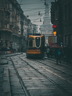 A tram is traveling down a wet cobblestone street in a city with overhead power lines and historic buildings lining the sides. There is a crowd of people on the right, some holding umbrellas, suggesting it might be raining or has recently rained. Neon signs, including one for a kebab and pizza shop, add touches of color to the predominantly gray and somber scene.