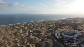 Aerial view of vast sand dunes stretching towards the horizon with a backdrop of ocean waves gently meeting the shore. In the foreground, a cluster of white buildings with lush greenery appears to be part of a resort or residential area. The sky is partly cloudy with soft lighting indicating the time might be early morning or late afternoon.