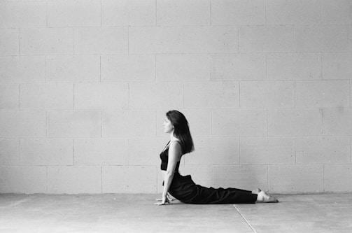 A person in a seated yoga pose on the floor next to a large, plain brick wall. The setting is minimalistic with a focus on the calm and composed posture of the individual.