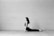 A person in a seated yoga pose on the floor next to a large, plain brick wall. The setting is minimalistic with a focus on the calm and composed posture of the individual.
