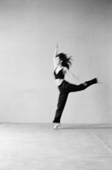 A blurred image features a person in motion, possibly dancing or performing, captured in mid-air with one arm raised and a leg extended. The background is a plain, textured wall, giving a minimalistic and artistic feel.