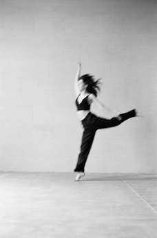 A blurred image features a person in motion, possibly dancing or performing, captured in mid-air with one arm raised and a leg extended. The background is a plain, textured wall, giving a minimalistic and artistic feel.
