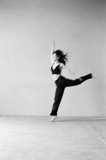 A blurred image features a person in motion, possibly dancing or performing, captured in mid-air with one arm raised and a leg extended. The background is a plain, textured wall, giving a minimalistic and artistic feel.