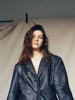 A person with long brown hair is wearing a dark leather jacket. The background consists of a plain, neutral-colored drape.