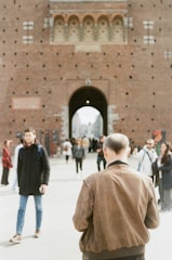 People walk near a historic brick castle with a prominent archway. The scene includes individuals dressed in casual attire and the focus is on a man wearing a brown jacket, seen from behind.