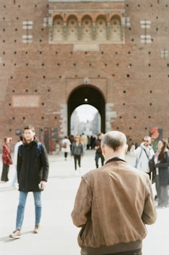 People walk near a historic brick castle with a prominent archway. The scene includes individuals dressed in casual attire and the focus is on a man wearing a brown jacket, seen from behind.