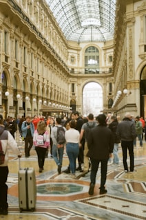 A large, elegant, and ornate shopping arcade with a glass-domed roof, featuring intricately detailed architecture and patterns on the floor. The area is bustling with people walking and interacting, adding a lively atmosphere.