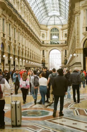 A large, elegant, and ornate shopping arcade with a glass-domed roof, featuring intricately detailed architecture and patterns on the floor. The area is bustling with people walking and interacting, adding a lively atmosphere.
