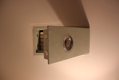 A wall-mounted safe with a metallic combination dial, partially opened to show its empty interior. The safe is embedded in a light-colored wall, and it is constructed from a textured material.