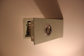A wall-mounted safe with a metallic combination dial, partially opened to show its empty interior. The safe is embedded in a light-colored wall, and it is constructed from a textured material.