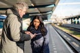 Two people stand on a train station platform. One person is holding a map or mobile device, and the other is looking intently at it. The platform has a modern design with a covered roof and train tracks running parallel.