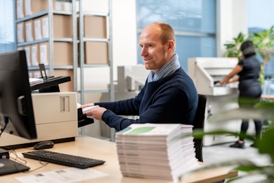A man is sitting at a desk, engaged in using a printer in a modern office setting. Shelves with boxes are visible in the background, and a stack of papers is on the table. Behind him, another person is operating a large copy machine. Indoor plants are visible near the window, giving a sense of a well-organized, professional workspace.