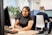 A woman wearing a headset sits at a desk, focused on her computer screen while taking notes with a pen. She is in a modern office setting with other people in the background also wearing headsets and working at computers. Plants and office partitions are visible.