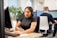 A woman wearing a headset sits at a desk, focused on her computer screen while taking notes with a pen. She is in a modern office setting with other people in the background also wearing headsets and working at computers. Plants and office partitions are visible.