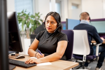A woman wearing a headset sits at a desk, focused on her computer screen while taking notes with a pen. She is in a modern office setting with other people in the background also wearing headsets and working at computers. Plants and office partitions are visible.