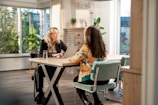 Two women are sitting at a wooden table in a well-lit room with large windows. One is using a laptop and the other is attentively looking on. The room is decorated with indoor plants and wooden furniture, giving it a modern, cozy feel.