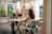 Two women are sitting at a wooden table in a well-lit room with large windows. One is using a laptop and the other is attentively looking on. The room is decorated with indoor plants and wooden furniture, giving it a modern, cozy feel.