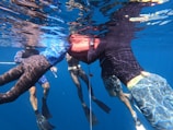 Four people underwater appear to be engaging in an activity with a red float. They are wearing snorkels and fins, suggesting this is a diving or snorkeling activity. The water around them is clear, and there are reflections on the surface.