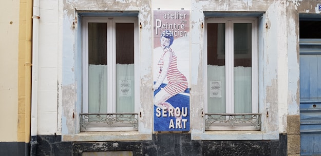 A building facade features two windows with white framed glass and metal grills underneath. Between them, there's a vertical sign with an image of a person wearing striped clothing on a blue background. The text on the sign includes 'atelier, Peintre, SEROU ART'. The surrounding wall is weathered with patches of peeling paint.