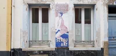 A building facade features two windows with white framed glass and metal grills underneath. Between them, there's a vertical sign with an image of a person wearing striped clothing on a blue background. The text on the sign includes 'atelier, Peintre, SEROU ART'. The surrounding wall is weathered with patches of peeling paint.