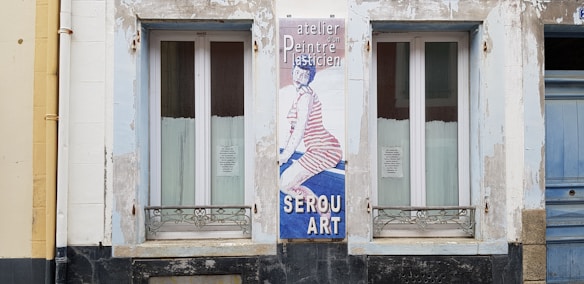A building facade features two windows with white framed glass and metal grills underneath. Between them, there's a vertical sign with an image of a person wearing striped clothing on a blue background. The text on the sign includes 'atelier, Peintre, SEROU ART'. The surrounding wall is weathered with patches of peeling paint.