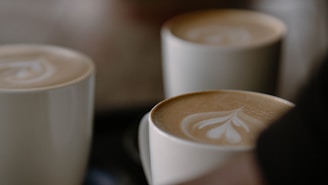 Several cups of coffee with intricate latte art on the surface, featuring heart and leaf designs in a creamy foam. The cups are arranged closely together, casting soft shadows.