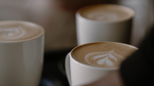 Several cups of coffee with intricate latte art on the surface, featuring heart and leaf designs in a creamy foam. The cups are arranged closely together, casting soft shadows.