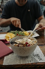 A person is sitting at a table preparing to eat, with a bowl of granola, berries, and yogurt in the foreground. Nearby, there is a plate with an omelette and some greens. The table is set with a red napkin and the bowl rests on a piece of paper resembling newspaper.