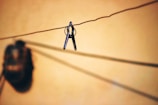A pair of scissors hangs from a wire against a warm, blurred background. The shadow of a round object is visible nearby, creating a contrast with the sharp focus of the scissors.