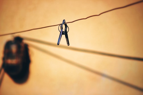 A pair of scissors hangs from a wire against a warm, blurred background. The shadow of a round object is visible nearby, creating a contrast with the sharp focus of the scissors.