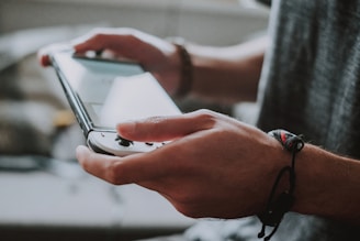 A person holds a handheld gaming console with both hands, with the screen illuminated. The individual wears a bracelet on their wrist, and the focus is on the device and hands, not revealing the person's face.