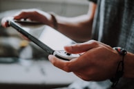 A person holding a handheld gaming console, focusing on the screen. The scene suggests an interactive moment, likely involving a video game. The person wears a bracelet on their right wrist. Natural lighting creates a soft ambiance.