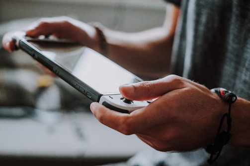 A person holding a handheld gaming console, focusing on the screen. The scene suggests an interactive moment, likely involving a video game. The person wears a bracelet on their right wrist. Natural lighting creates a soft ambiance.